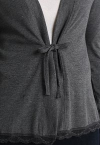 Mörkgrå ribbad cardigan knuten framtill med rosett, med spetskant vid fållen, bärs över en vit topp.