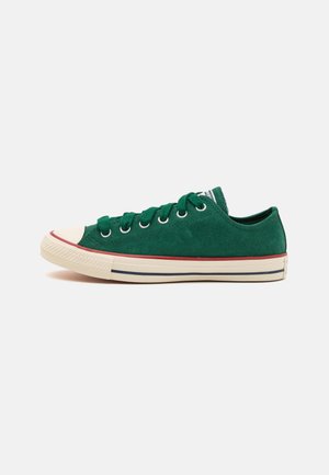 Zapatilla baja de ante verde con punta redonda, suela de goma crema en contraste y detalle de raya distintiva. Seis ojales con cordones verdes.