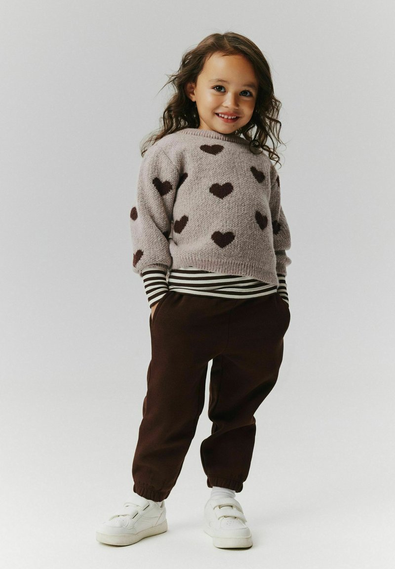 Jeune fille souriante aux cheveux bouclés foncés portant un pull à motif cœur, une chemise rayée, un pantalon foncé, des chaussettes blanches et des baskets blanches.