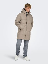 Beige puffer coat with a hood, featuring vertical quilted design, two front pockets, and black zipper accents. Worn with light blue jeans and white sneakers.