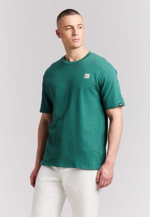 Man met kort haar die een groen shirt met korte mouwen en een witte broek draagt, kijkt naar links tegen een effen lichte achtergrond.
