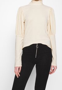 Vaalean beige, raidallinen pitkähihaisia paita, jossa on korkea kaulus ja puffattuja hartioita, yhdistettynä mustiin pystysuorasti raidoitettuihin housuihin, joissa on vetoketjullinen etuosa.