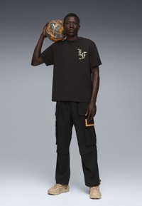 Sort bomulds-T-shirt med guld-logo, parret med sorte cargo-bukser med orange detaljer. Modellen holder en tekstureret orange og grå basketball.