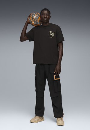 Sort bomulds-T-shirt med guld-logo, parret med sorte cargo-bukser med orange detaljer. Modellen holder en tekstureret orange og grå basketball.