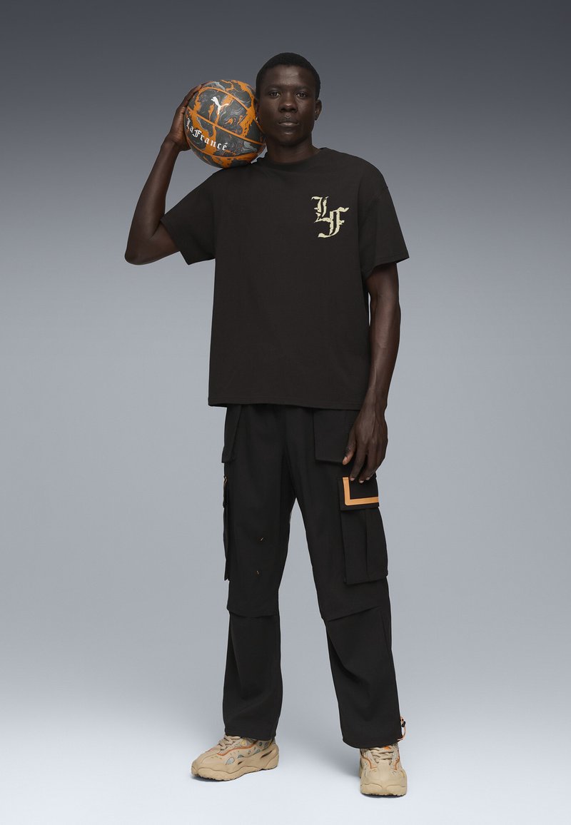 Sort bomulds-T-shirt med guld-logo, parret med sorte cargo-bukser med orange detaljer. Modellen holder en tekstureret orange og grå basketball.