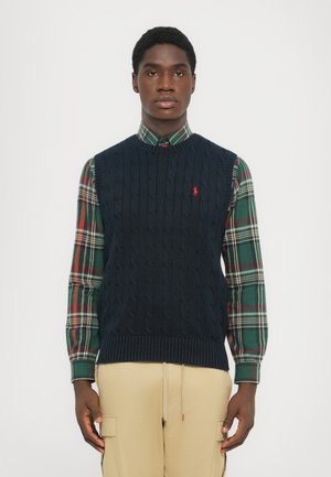 Hombre que lleva un chaleco de punto en azul marino sobre una camisa de cuadros verde y roja y pantalones cargo beige, de pie contra un fondo liso.