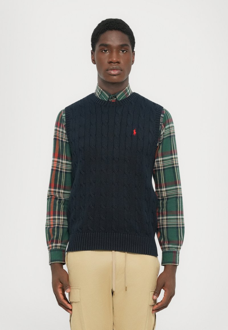 Hombre que lleva un chaleco de punto en azul marino sobre una camisa de cuadros verde y roja y pantalones cargo beige, de pie contra un fondo liso.