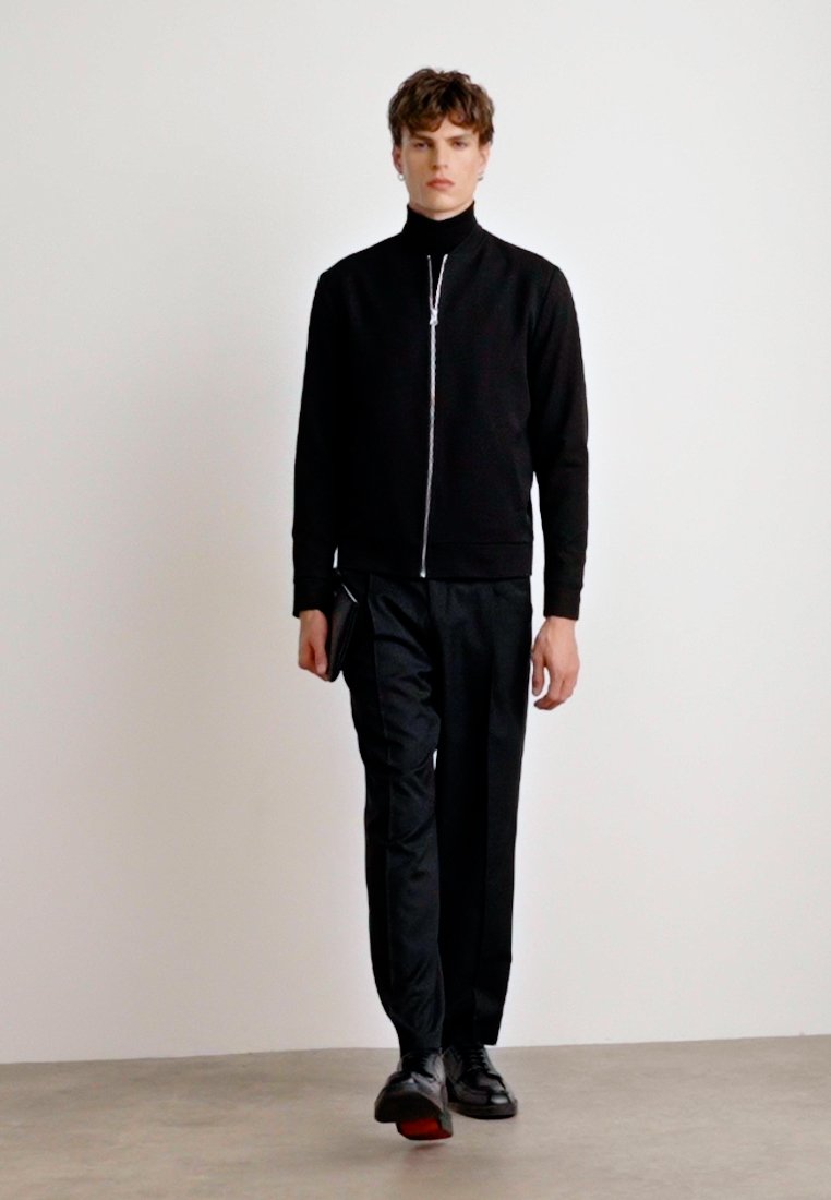 Veste noire zippée avec un col montant, associée à un pantalon noir. L'ensemble est complété par des chaussures noires brillantes et un accessoire discret au poignet.