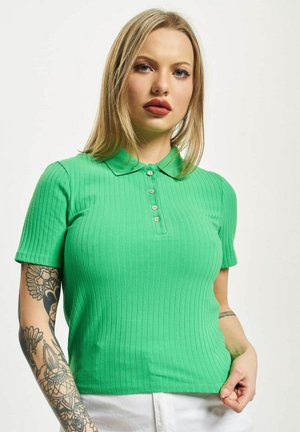 Eine blonde Frau mit leichtem Make-up trägt ein hellgrünes, geripptes Poloshirt, weiße Hosen und hat Blumen-Tattoos am linken Unterarm.