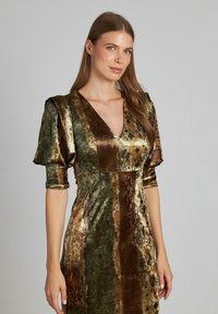 Una donna con lunghi capelli castani indossa un abito di velluto nei toni del verde e del marrone, con maniche a sbuffo e scollatura a V, su uno sfondo grigio uniforme.