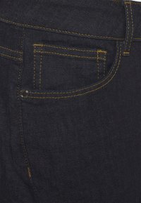 Pantalones vaqueros de mezclilla azul oscuro con costuras amarillas, con un bolsillo trasero y detalle de remache de metal. Textura suave, corte clásico.