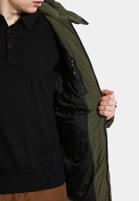 Veste matelassée vert olive avec un intérieur noir, comportant une poche zippée et une doublure texturée. Le modèle tient la veste ouverte.