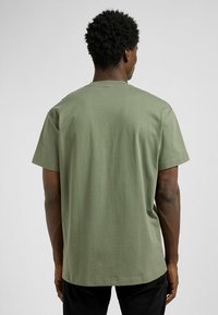 Oliwkowa bawełniana koszulka z luźnym krojem, krótkimi rękawami i okrągłym dekoltem. Gładka tekstura, prosty design, brak widocznych wzorów lub grafik.