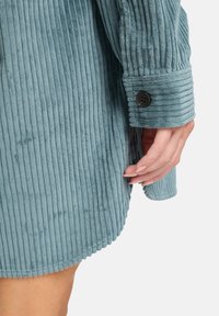 Tegenblauwe corduroy shirt met verticale ribbels, voorzien van een afgeronde zoom en knoopmanchetten. Zachte textuur en duidelijke stikdetails.