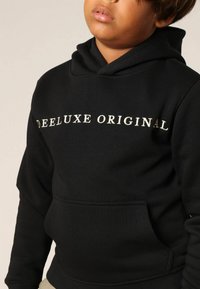 Zwarte hoodie gemaakt van zachte stof, met een voorkantzak en de tekst "DEE LUXE ORIGINAL" in crème. Regular fit met een capuchon met trekkoord.