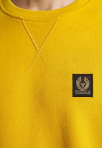 Gul bomullströja med rund halsringning, korsstygnsdetalj och en svart logotypapplikation på vänster bröst. Mjuk textur genomgående.