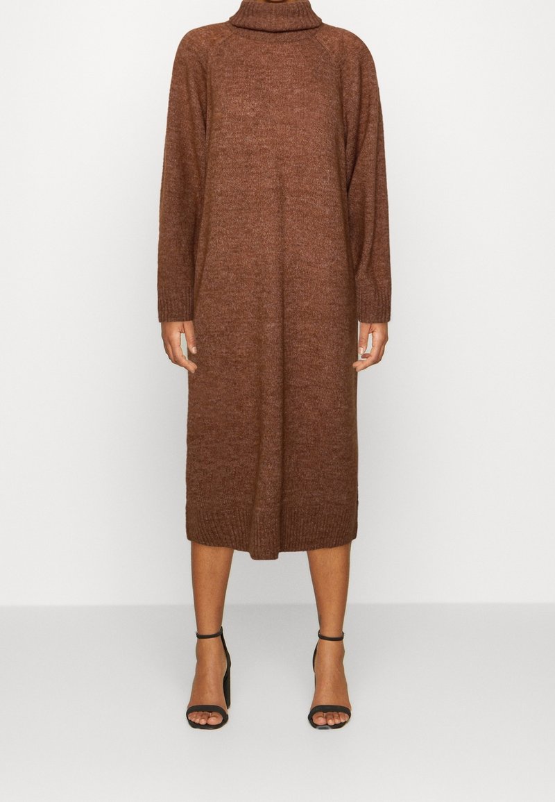 Robe midi en maille à col roulé marron à manches longues portée avec des sandales noires à talons hauts et à bride à la cheville, modèle debout sur un fond clair uni.