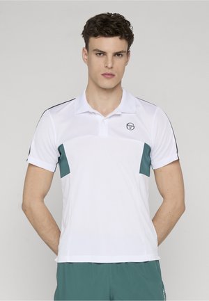 Ung mand med mørkt hår, iført en hvid polo med grønne sidepaneler og grønne shorts, stående mod en ensfarvet baggrund.