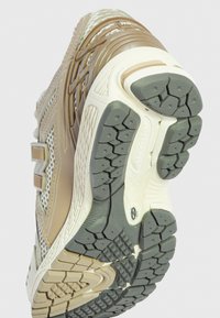 Chaussure de course avec une tige en mesh beige, une semelle en caoutchouc grise, des accents marron texturés et des rainures flexibles pour une meilleure traction.