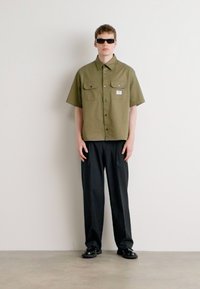 Short-sleeve olive green collared shirt with two chest pockets, paired with black trousers and black shoes, worn with dark sunglasses.