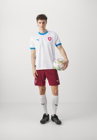 Balts futbols krekls ar ziliem akcentiem un emblēmu, sarkanas šortus, melnas sporta kurpes un baltas zeķes. Spēlētājs tur krāsainu futbolu.