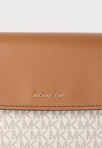 Ταμπά δερμάτινο καπάκι με χρυσό λογότυπο "MICHAEL KORS" πάνω από κρεμ ύφασμα με επαναλαμβανόμενο μοτίβο "MK" σε ταμπά χρώμα. Επιφάνεια με υφή.