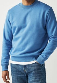 Sweat-shirt bleu clair en matière douce, avec un col rond côtelé et des poignets, associé à un jean bleu. Texture lisse, coupe décontractée.