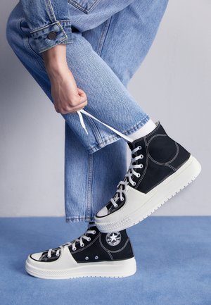 Person iført lyseblå jeans og sorte høje sneakers med hvide såler binder en skosnøre på en blå gulv med en lys grå baggrund.