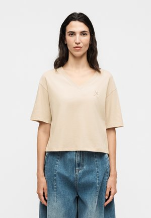 Kvinde med mørkt hår iført en beige kortærmet V-hals skjorte og højtaljede blå denimjeans, stående mod en ensfarvet baggrund.