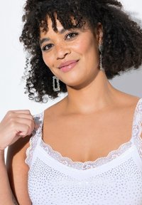 Top bianco con bordo in pizzo e decorazioni in strass. Presenta una scollatura tonda e spalline sottili, mettendo in evidenza una texture di tessuto liscia.