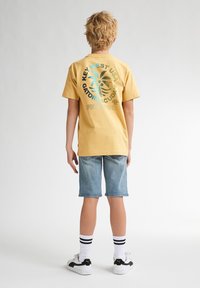 Gul bomulds-T-shirt med et stort grafisk print i blå og grøn på ryggen, parret med lyseblå denimshorts og sort-hvide sneakers.