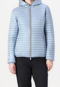 Lyseblå pufferjakke med lynlås foran, horisontal quiltet tekstur, fast hætte og elastiske manchetter, parret med sorte bukser.