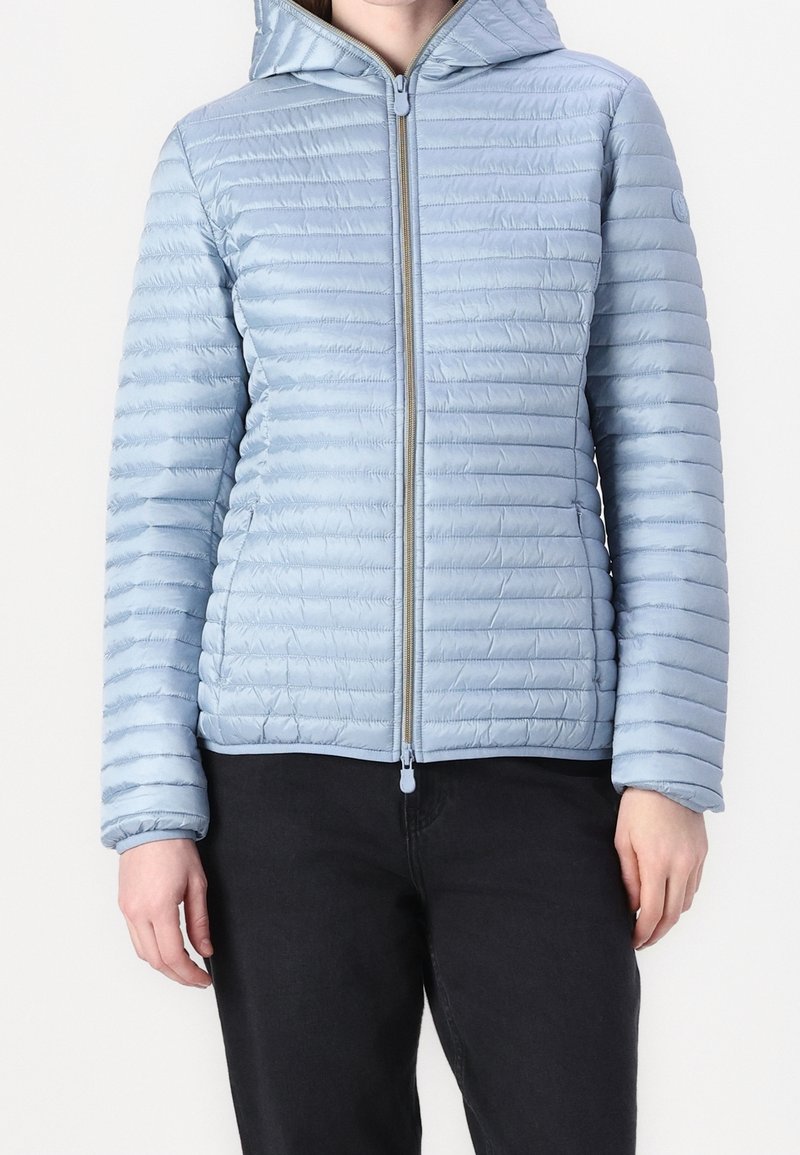 Lyseblå pufferjakke med lynlås foran, horisontal quiltet tekstur, fast hætte og elastiske manchetter, parret med sorte bukser.