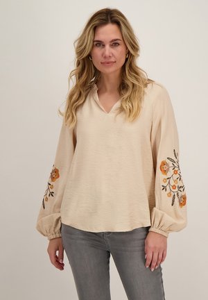 Vrouw met een beige blouse met oranje bloemrijke borduursels op ballonmouwen en grijze jeans, staand tegen een egale achtergrond.
