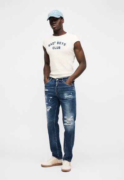Jeune homme portant un débardeur blanc avec l'inscription "BAD² BOYS CLUB", un jean bleu déchiré, des baskets blanches et une casquette de baseball bleu clair, debout les mains dans les poches.
