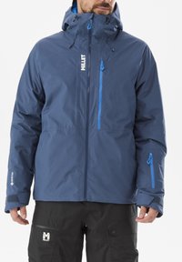 Man wearing a blue waterproof hooded jacket and black pants with brand logos, standing against a white background.