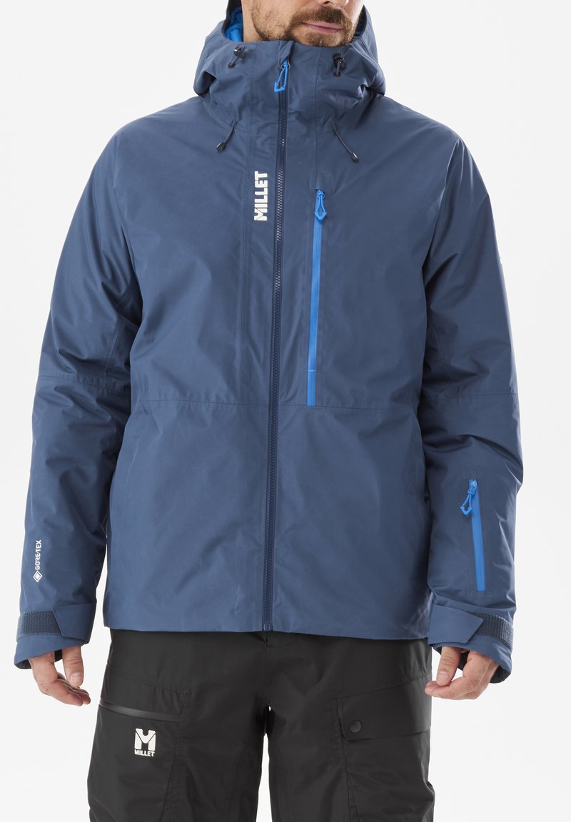 Man wearing a blue waterproof hooded jacket and black pants with brand logos, standing against a white background.