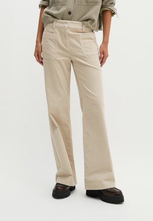 Personne portant un pantalon beige à jambes larges avec poches avant et des chaussures marron à semelles épaisses, debout devant un fond uni.