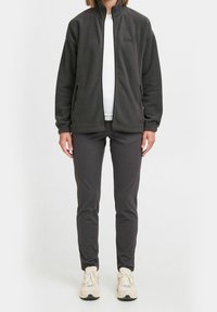 Grå fleecejakke med høj krave, lynlåslommer og logo på brystet, kombineret med mørke bukser og lyse sneakers.