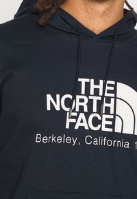 Osoba v tmavé mikině s kapucí, na které je bílé logo "The North Face" a text "Berkeley, California" na hrudi, viditelné tkanice mikiny.