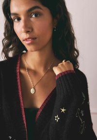 Cárdigan negro con ribetes rojos, que presenta bordados dorados de estrellas y un diseño. El modelo lleva un collar dorado con un colgante texturizado.
