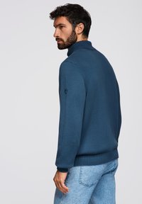Dunkelblauer Strickpullover mit hohem Kragen, gerippten Bündchen und einem strukturierten Muster. Mit einem kleinen Akzentlogo am Ärmel.