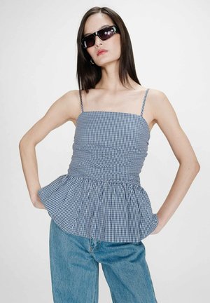 Femme portant des lunettes de soleil noires, un haut sans manches à carreaux bleu et blanc avec un volant à l'ourlet, et un jean bleu clair, posant les mains sur les hanches.