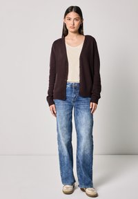 Mörkbrun cardigan i mjukt stickat material, öppen framsida med knappar, matchad med en ljus beige topp och ljusblå vida jeans. Avslappnade skor ingår.