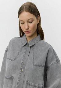 Giacca di jeans grigio chiaro con collo, due tasche frontali e bottoni a pressione argentati. La cucitura visibile contrasta con il tessuto.
