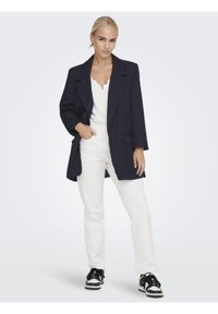 Blazer overdimensionato blu navy con revers a punta, abbinato a una maglietta bianca e pantaloni bianchi a vita alta. Sneakers bianche e nere completano il look.