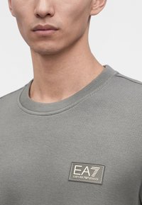 Homme portant un t-shirt gris texturé EA7 Emporio Armani à col rond avec écusson logo sur la poitrine gauche.