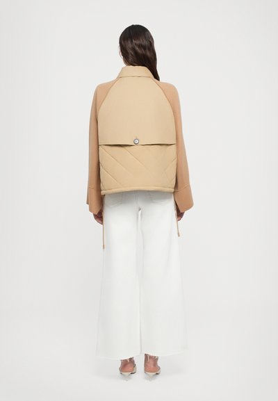 Tan quilted jacket with a flap, button closure, and drawstrings. Paired with white wide-leg pants and transparent heels.