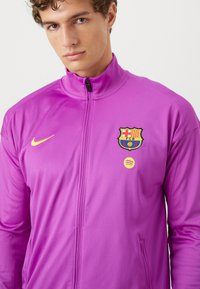 Paarse Nike-jack met volledige rits, staande kraag, Barcelona-embleem en geel Spotify-logo; gladde textuur, getailleerd ontwerp.