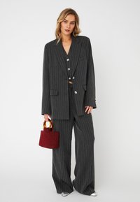 Abito grigio a righe sottili con pantaloni a gamba larga e un blazer corto, con bottoni lucidi; tiene una piccola borsa rossa testurizzata con manici in legno.